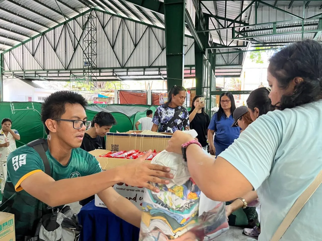 Two people pass a bag of donated goods across a table stacked with boxes inside a covered court, as others look on.