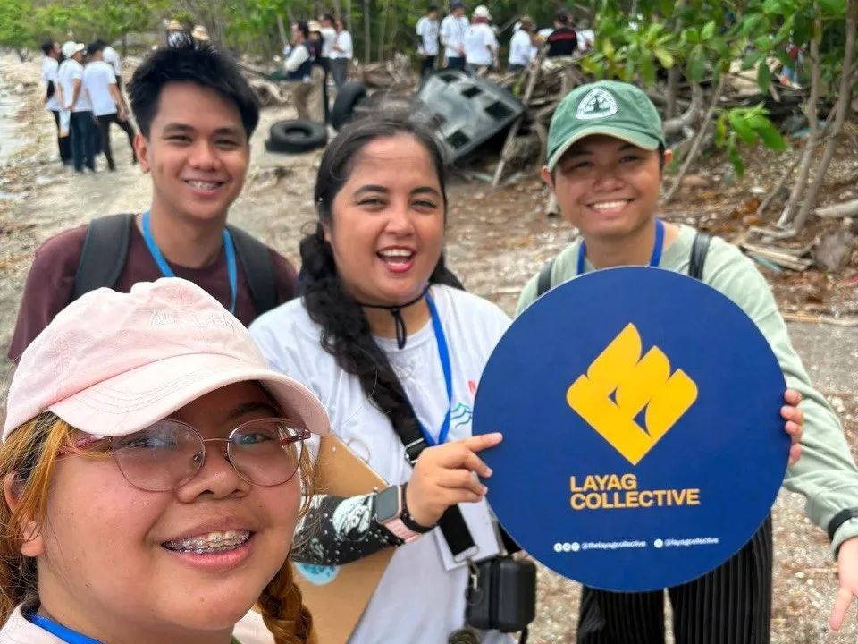 Four smiling volunteers take a selfie with a round Layag Collective sign at a coastal cleanup, as others clear debris behind them.